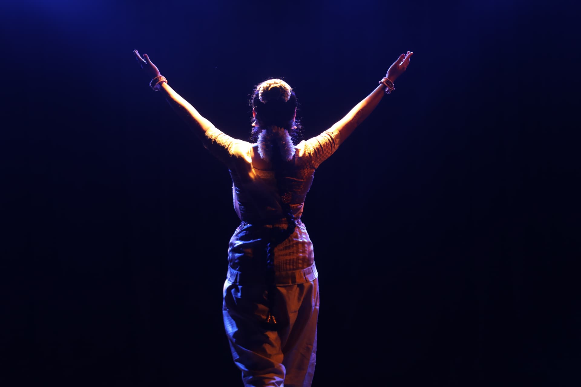 Bharatanatyam dancer raising both arms beneath blue stage lights